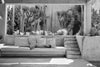 A black and white photograph of a person lying on a large outdoor lounge sofa with pillows, set in a sunlit architectural space with steps and desert plants in the background.