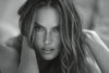 A close-up black and white portrait of a woman with long, wavy hair, looking directly at the camera with her hands framing her face.