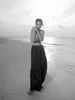 A black and white fashion photograph of a model standing on a sandy beach, wearing a dark, loose-fitting high-waisted maxi skirt and a matching dark top.