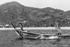 Black and white photograph of a longtail boat moving across water with two people aboard, with a sandy beach and forested mountains in the background.