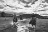 Black and white photograph of three cowboys riding horses through a muddy field, with mountains and a dramatic cloudy sky in the background.
